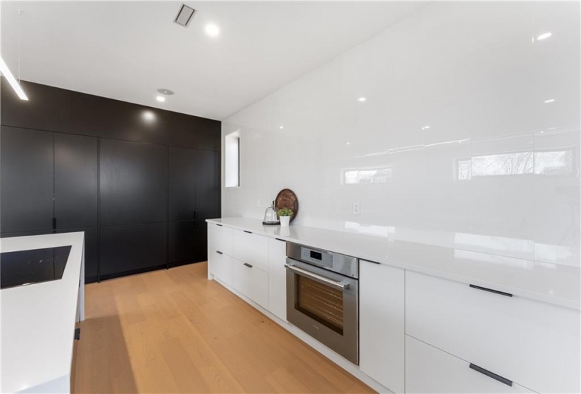 Integrated Thermador fridge. freezer, microwave, shelving (inside black cabinetry). White oak flooring. White quartz countertops. Multiple shelves, pull out drawers. Thermador cooktop, oven, dishwasher. Tiled white wall.